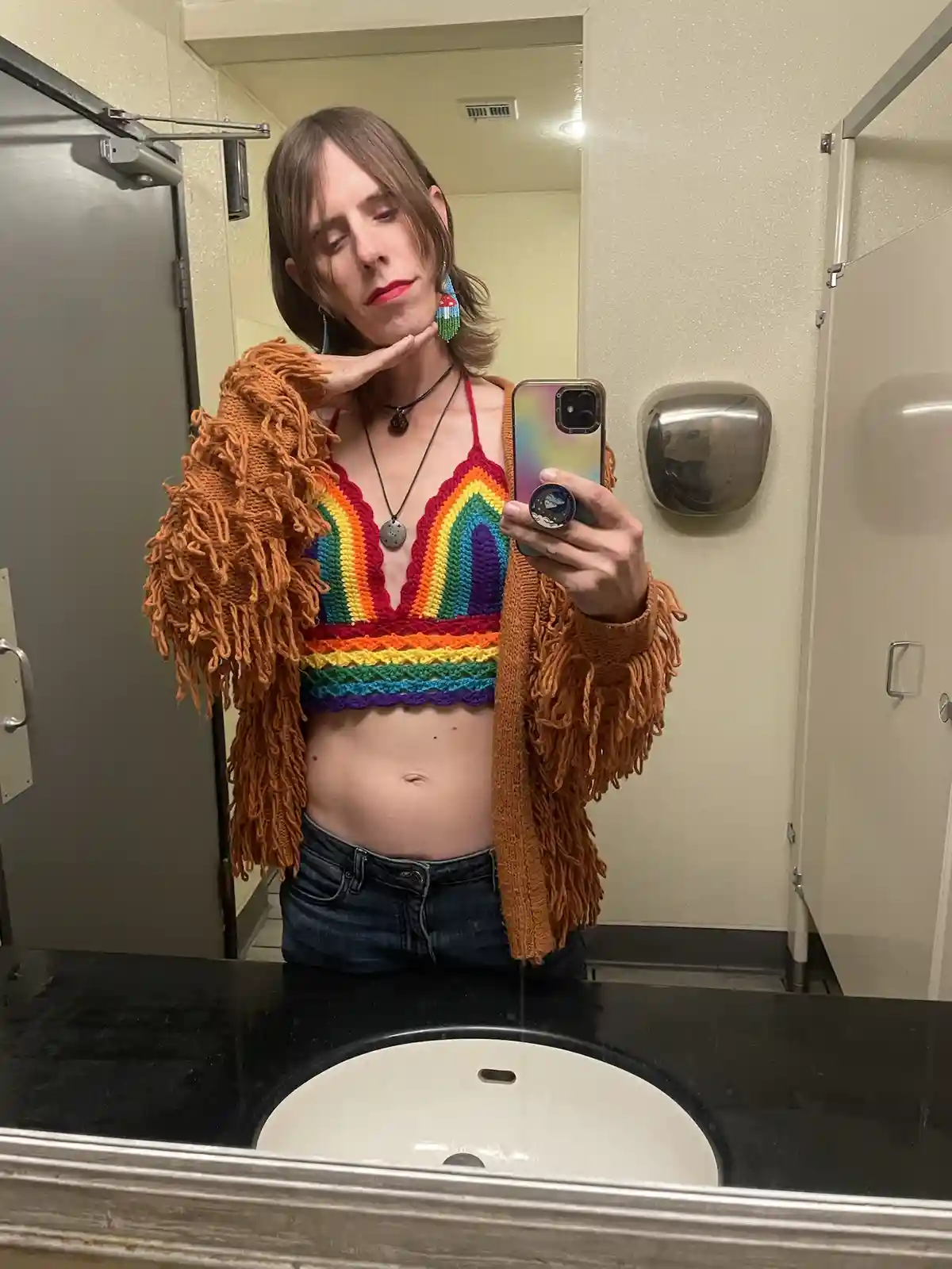 A person with shoulder-length brown hair and red lipstick takes a mirror selfie in a public restroom, wearing a rainbow…