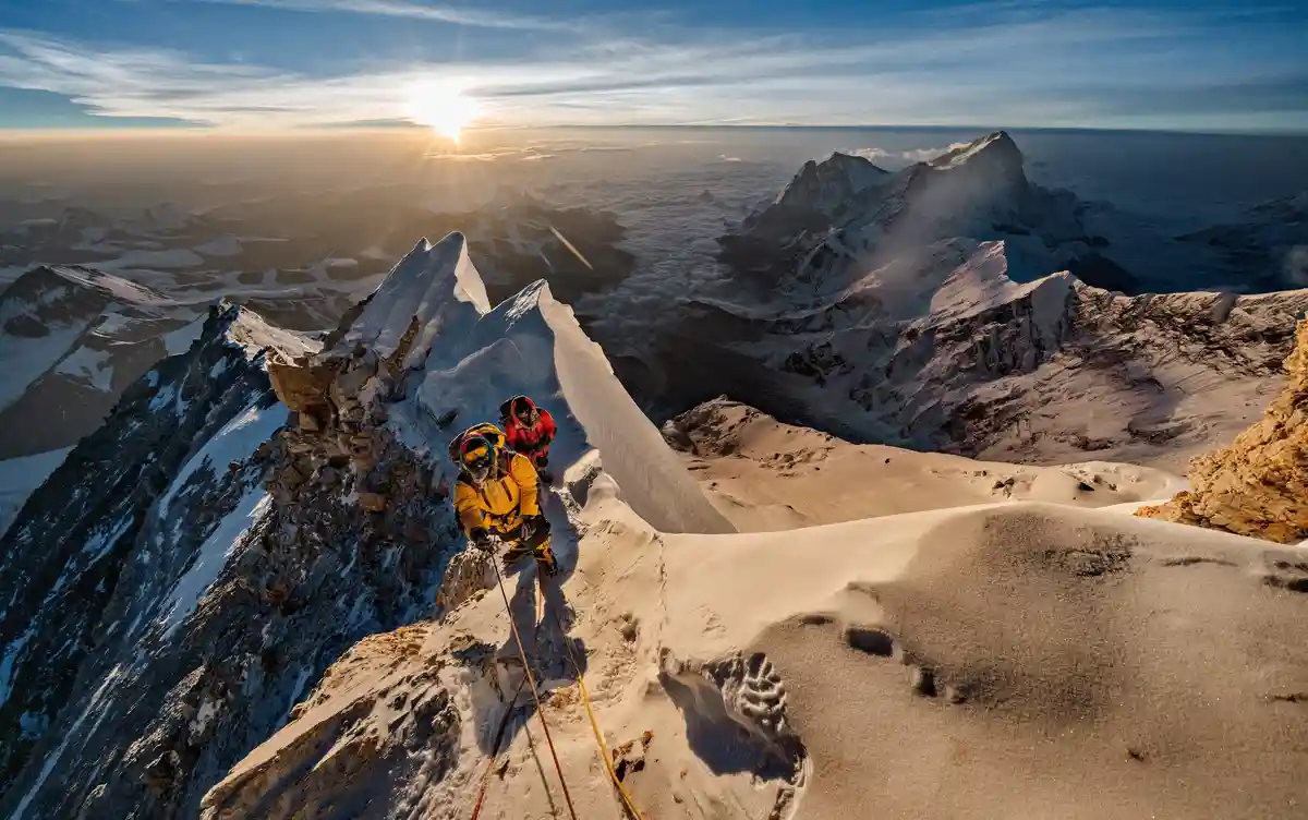 Two climbers in bright yellow and red gear ascend a steep, snow-covered mountain ridge under a sunrise, with a vast sea of…