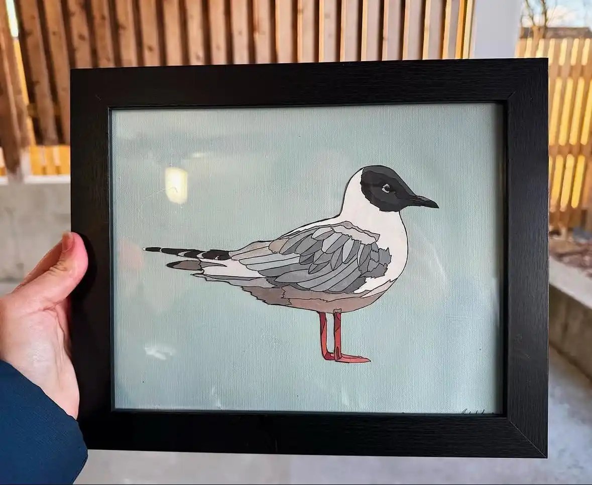 A hand holds a black-framed artwork depicting a stylized bird with a black head, white neck, gray wings, and red legs…