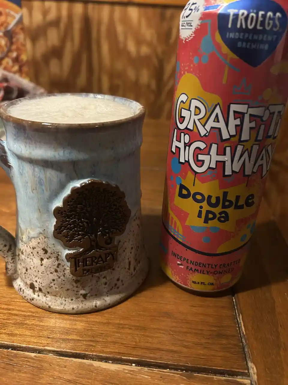 A ceramic mug with a tree logo and the words 'Therapy Brewing' sits next to a can of Troegs 'Graffiti Highway Double IPA'…