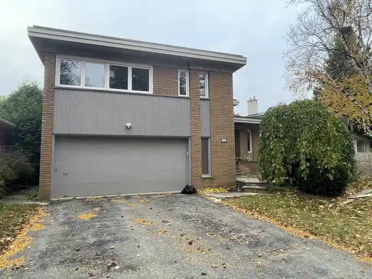 A two-story brick house with a large gray garage door and corrugated metal siding, surrounded by autumn leaves and bushes…