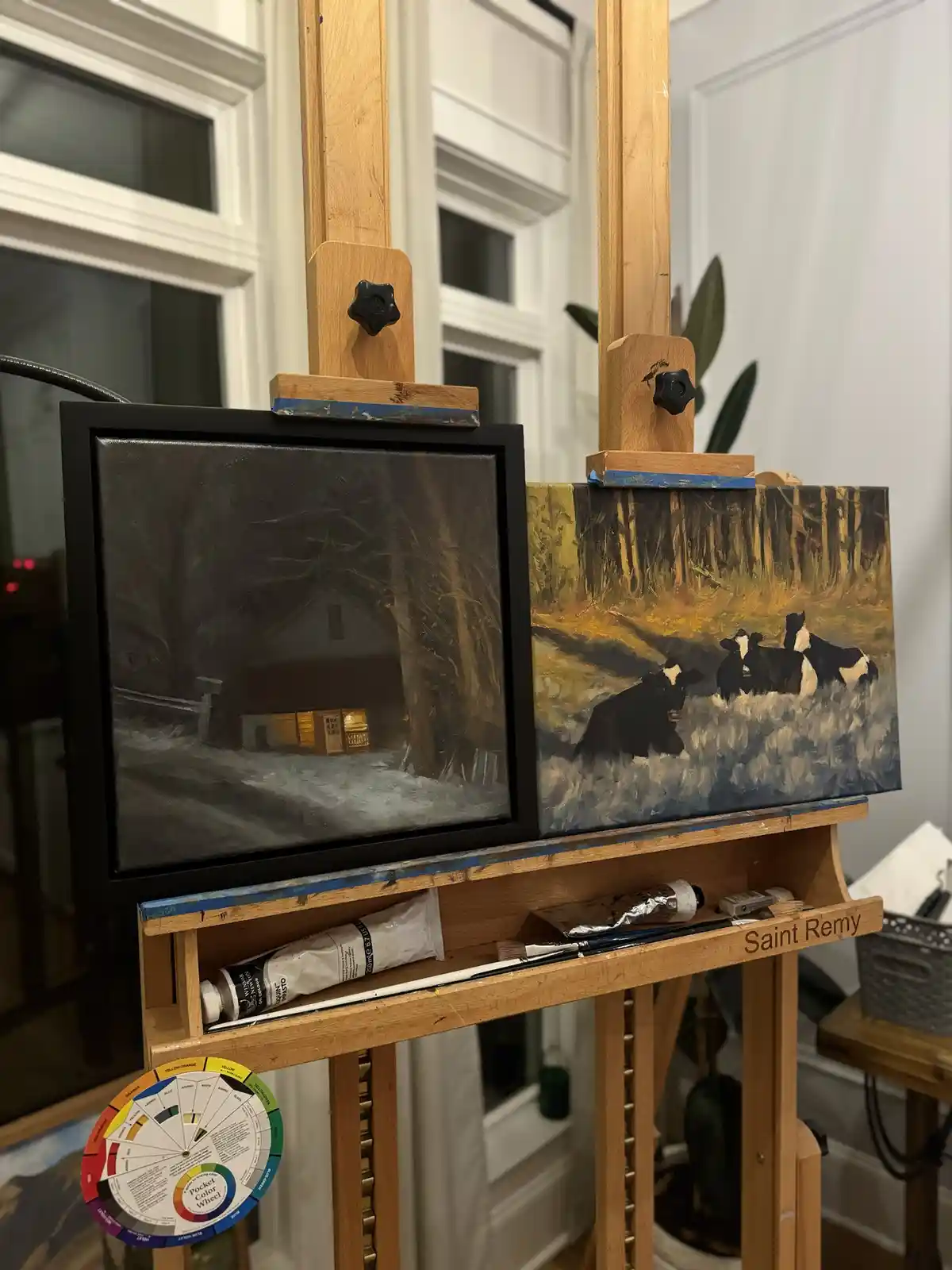 Two paintings sit side-by-side on a wooden easel: one depicts a dark, snowy scene with a lit house, the other shows cows in…
