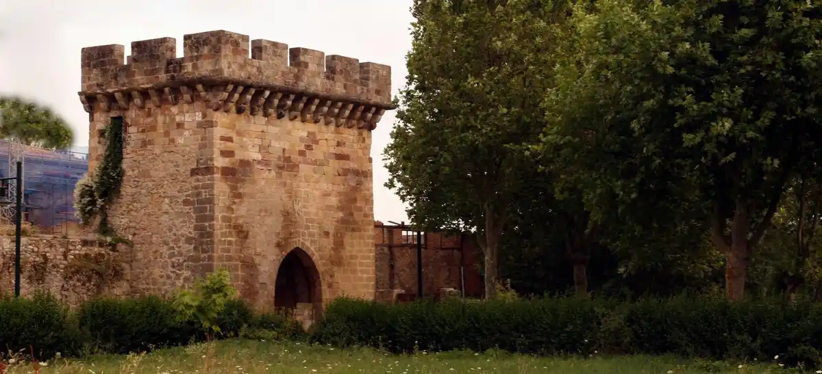 A stone castle tower with crenellations and an arched entrance, surrounded by green trees and bushes, with a modern blue…