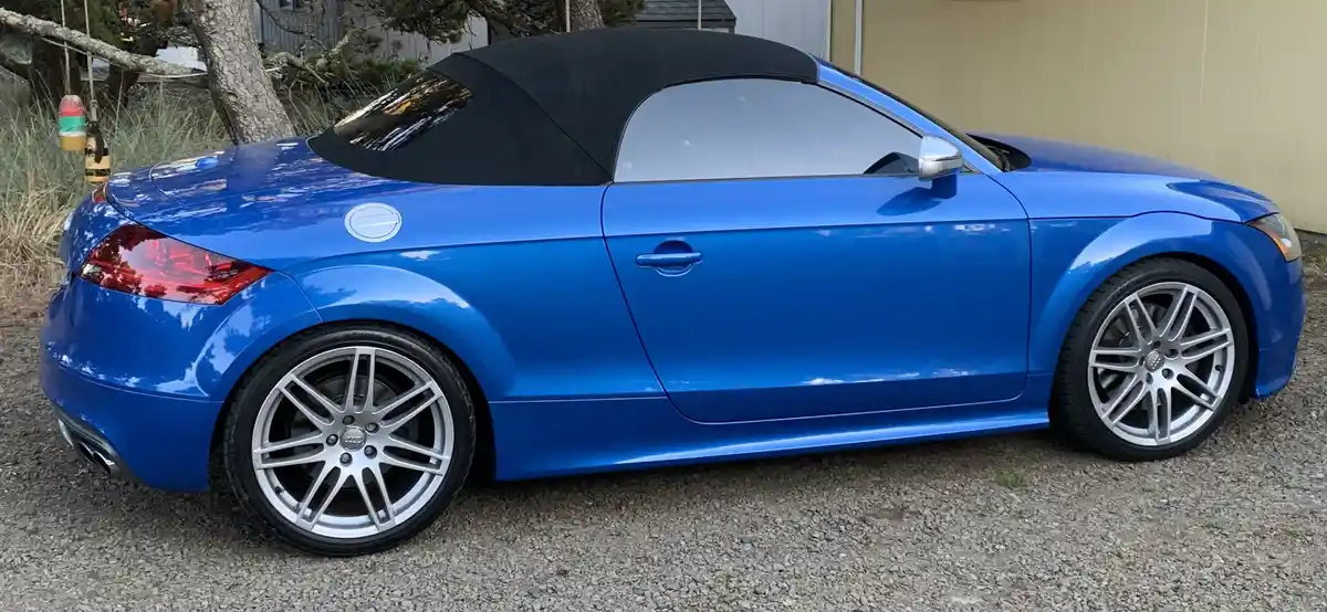 A shiny blue Audi TT roadster with a black soft top parked on gravel, featuring silver multi-spoke wheels and a sleek side…