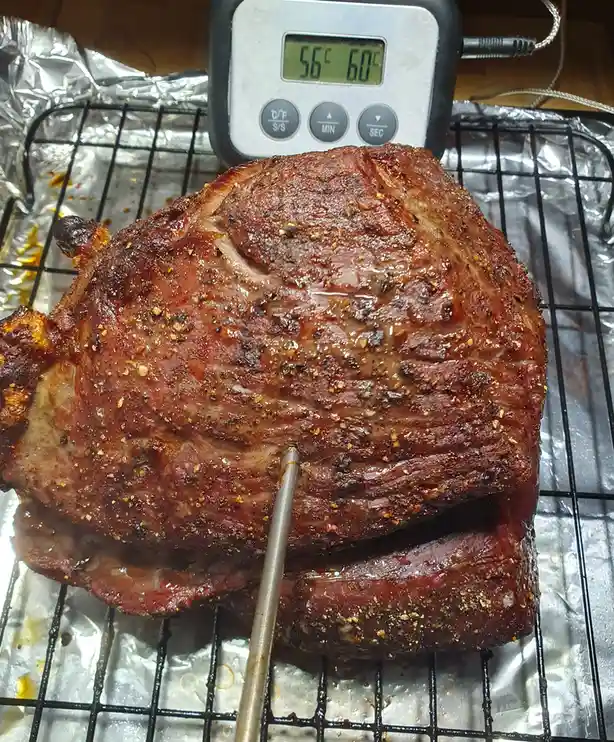 A large, seasoned, roasted meat roast sits on a wire rack over aluminum foil, with a digital meat thermometer inserted into…