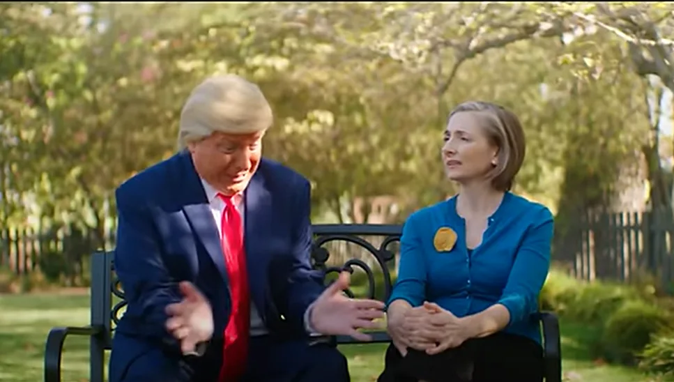 two people sit on a park bench; the man in a blue suit and red tie gestures animatedly while speaking, and the woman in a…
