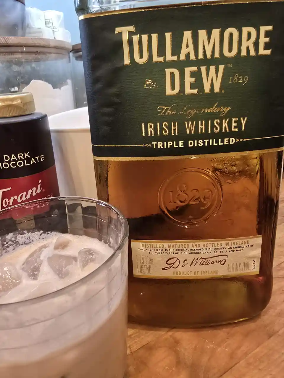 A bottle of Tullamore D.E.W. Irish whiskey sits next to a glass filled with ice and a creamy, frothy beverage, with a Torani…