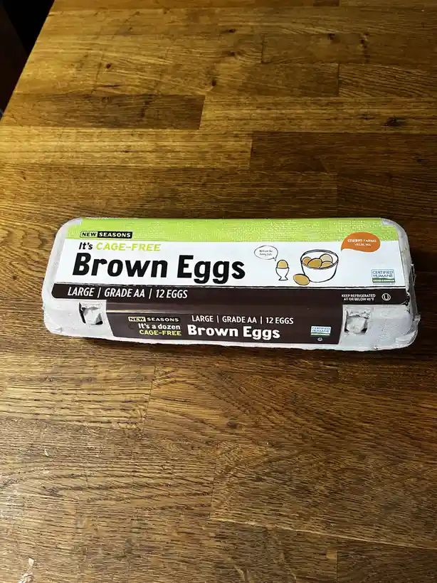 A carton of New Seasons brand brown eggs sits on a wooden table, with the label clearly stating 'It's a dozen cage-free…