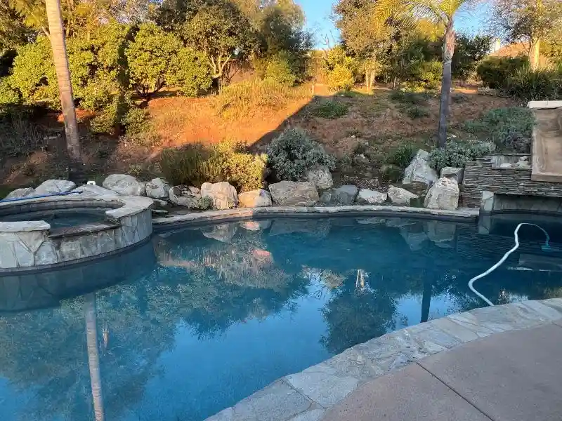 A serene outdoor swimming pool with clear blue water reflecting the sky and surrounding trees, bordered by stone tiles and…