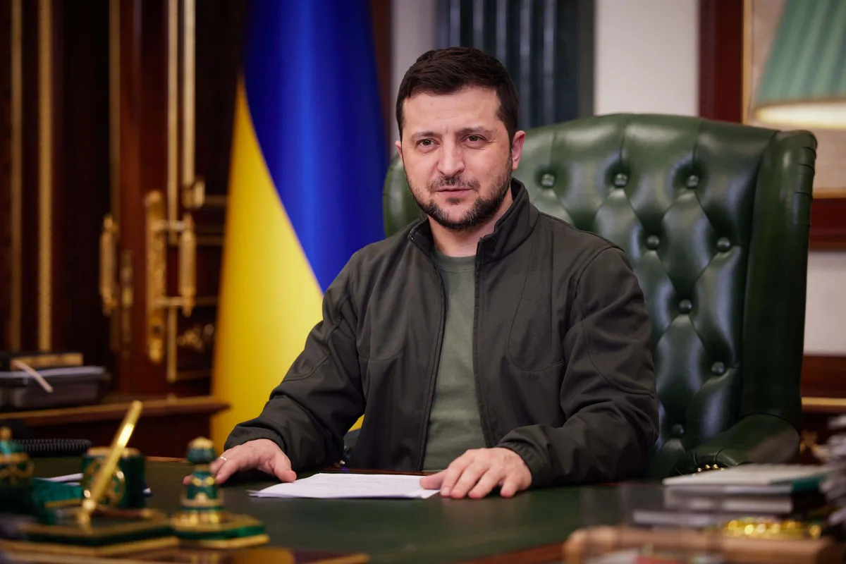 A man with a beard and short dark hair sits at a large green desk in an official-looking office, wearing a dark jacket over…