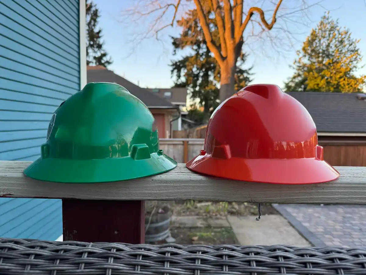 Two plastic construction helmets, one green and one red, sit side-by-side on a wooden railing outdoors. The green helmet has…