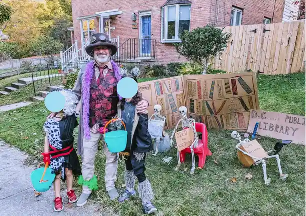 A family dressed in Halloween costumes stands in a front yard with cardboard signs and skeleton props. The man wears a…