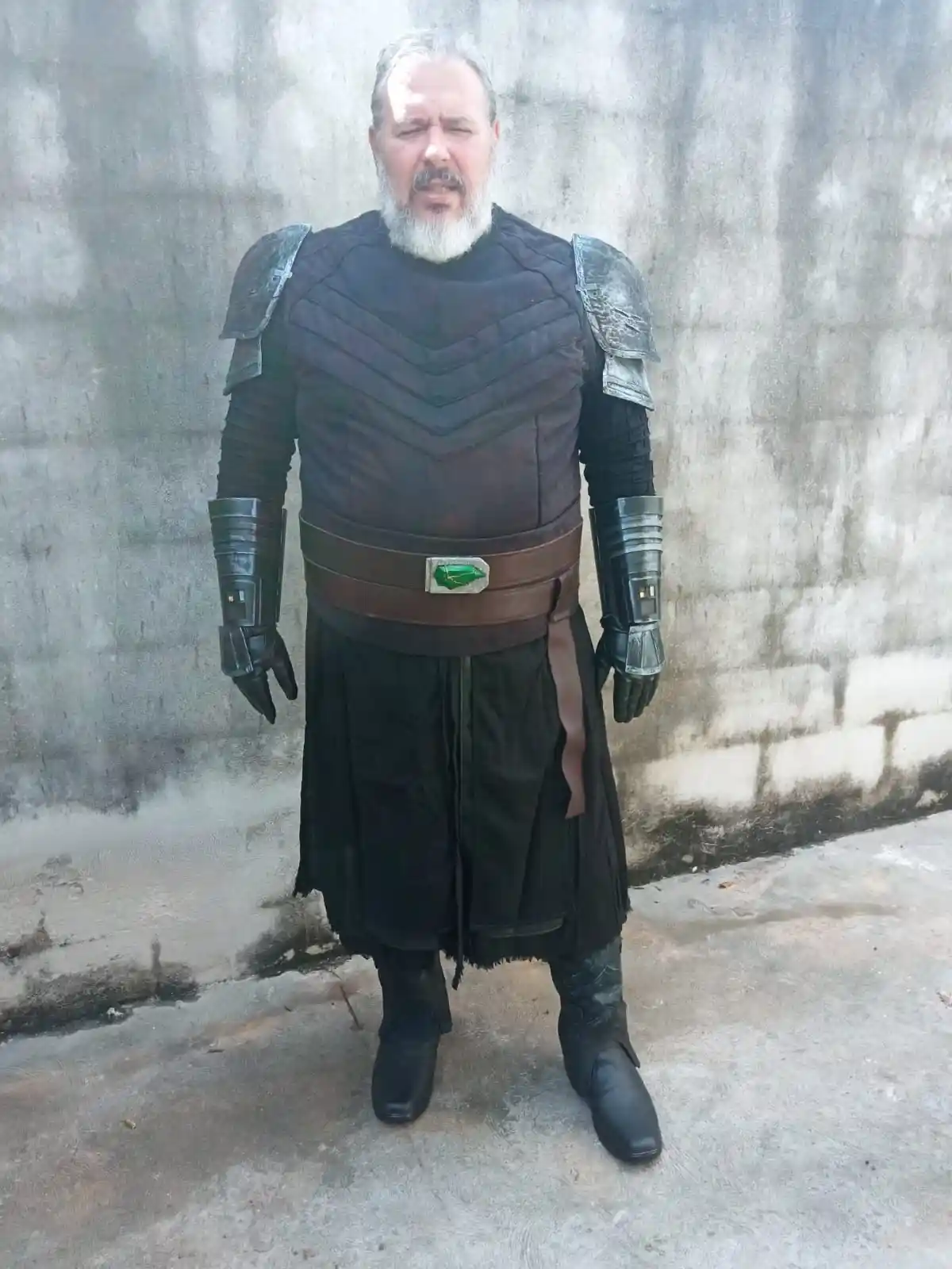 A man with a white beard and gray hair stands in front of a concrete wall wearing elaborate dark armor with shoulder pads…