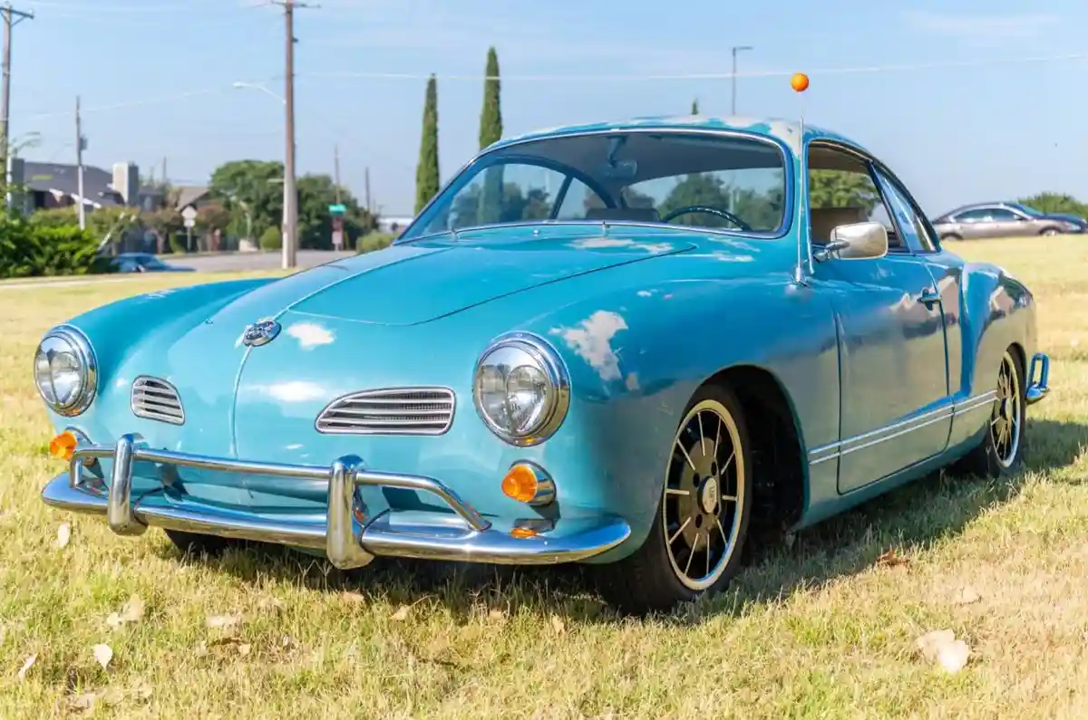 A vintage light blue Volkswagen Karmann Ghia is parked on dry grass under a clear sky, showing its classic rounded body…