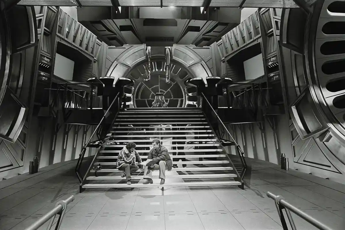 A black-and-white photograph shows two people sitting on wide metal stairs inside a large, futuristic spaceship set with…