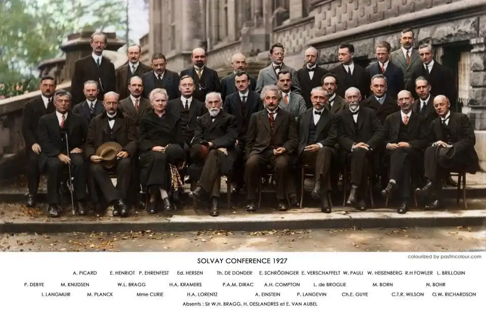 A colorized historical group photo from the 1927 Solvay Conference, showing dozens of prominent physicists in formal attire…