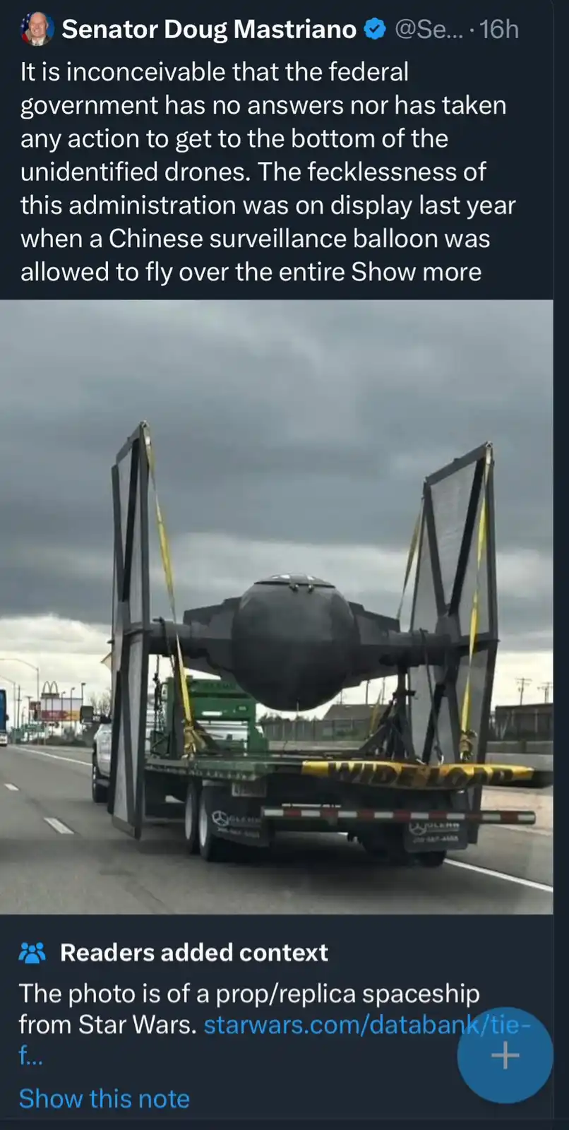 A green flatbed truck with a 'WIDE LOAD' sign is hauling a large, black, spherical object with two large, angular wings…