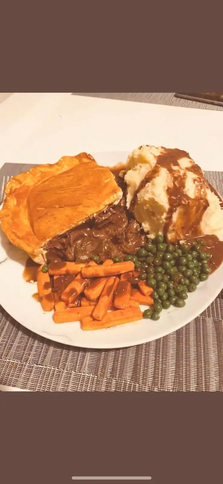 A plate of food featuring a partially eaten beef pie with golden crust, mashed potatoes with gravy, green peas, and carrots…