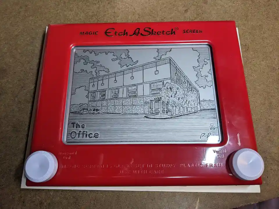 A red Etch A Sketch toy displays a detailed line drawing of the exterior of the TV show 'The Office' building, complete with…