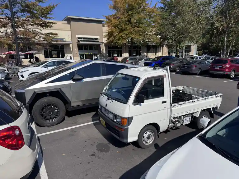 A silver, angular Tesla Cybertruck is parked next to a small, white Japanese mini truck in a sunny parking lot. The…