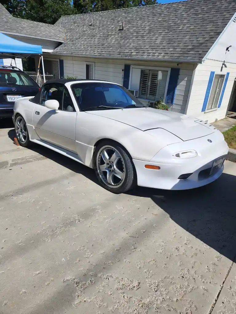 A white 1990 Mazda MX-5 Miata convertible is parked in a driveway in front of a white house with blue shutters. The car has…