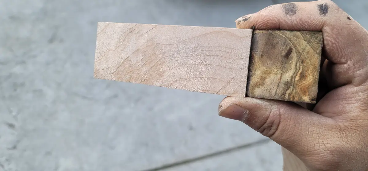 A hand holds a rectangular piece of wood with one end freshly sanded to a smooth, light-colored finish and the other end…