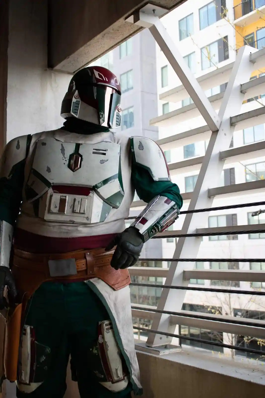 A person in a detailed Boba Fett costume stands on a balcony overlooking modern buildings, looking away from the camera. The…