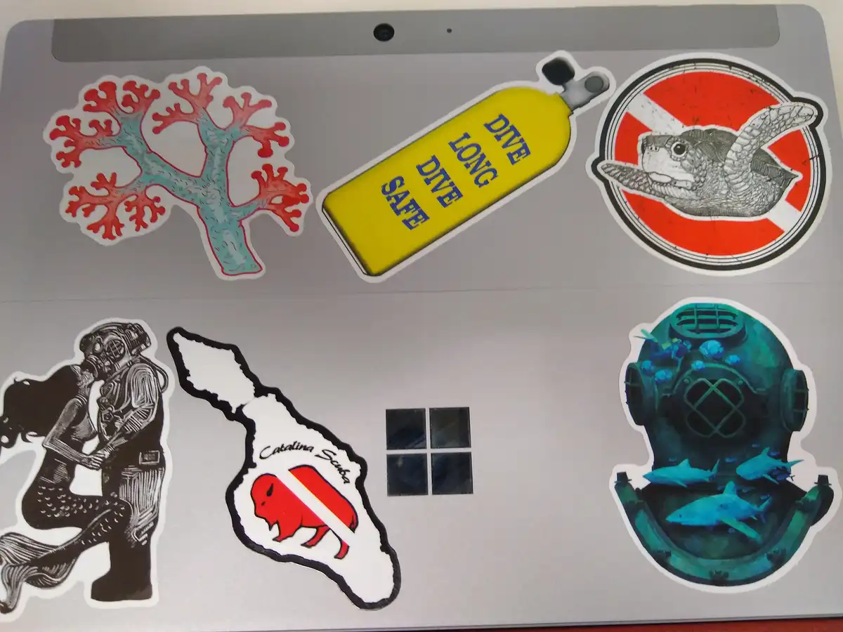 A laptop lid covered in dive-themed stickers including a coral, a yellow oxygen tank with 'DIVE LONG DIVE SAFE' text, a…