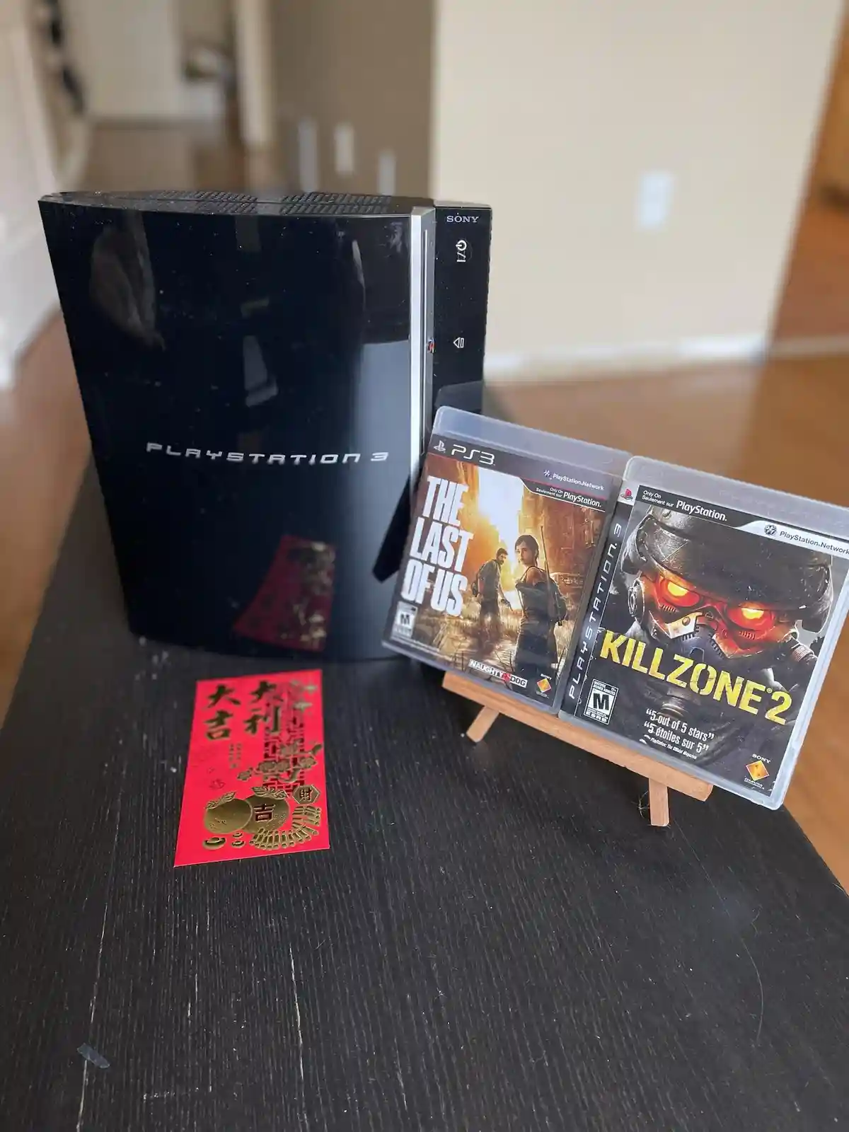 A black PlayStation 3 console sits on a dark wooden table next to two PS3 game cases for 'The Last of Us' and 'Killzone 2'…