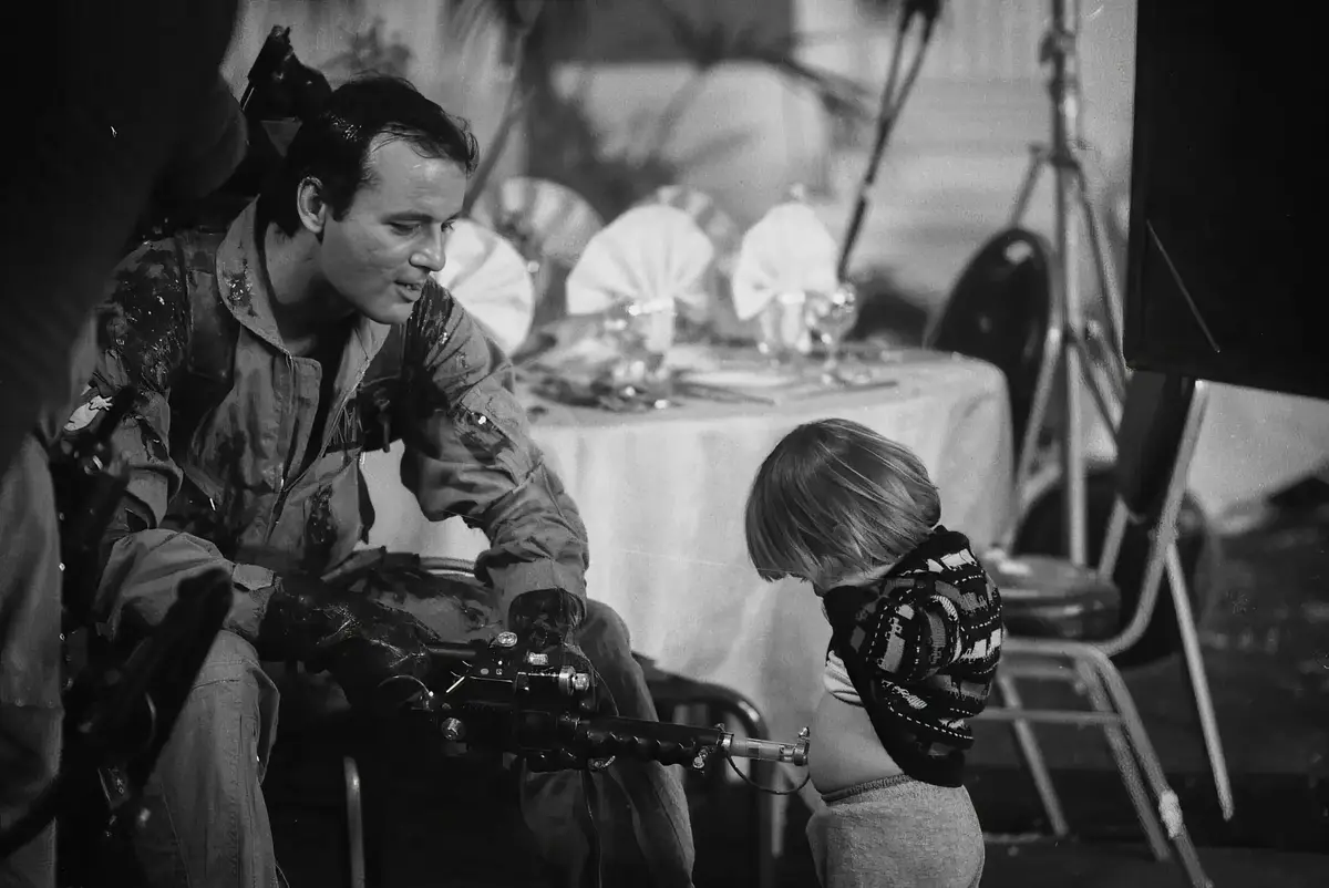 A black-and-white photo shows a man in a dirty, grease-stained jumpsuit holding a large, heavy machine gun, looking down at…
