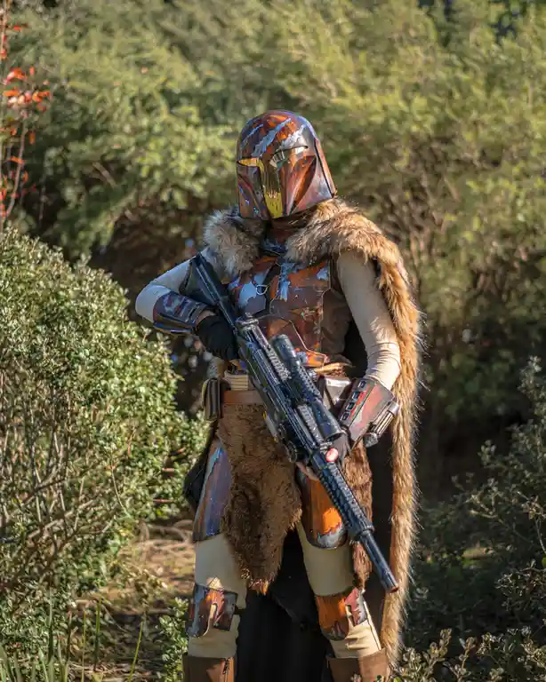 A person in a detailed, weathered Mandalorian armor costume with a fur-lined cape and helmet stands outdoors in a wooded…