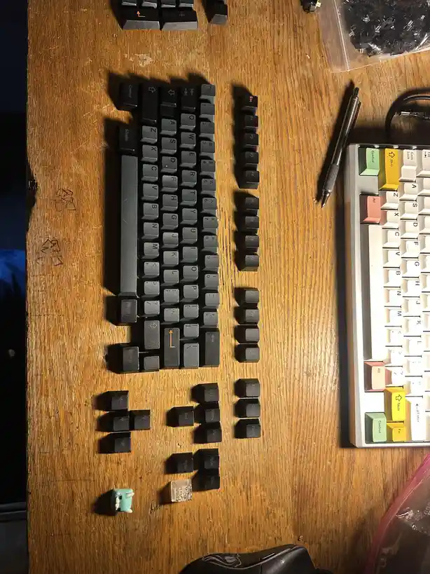 An overhead shot of a wooden desk with a partially disassembled black mechanical keyboard, its keys scattered around it. To…