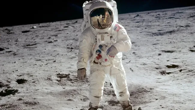 An astronaut in a white spacesuit stands on the moon's gray, cratered surface, helmet reflecting the lunar landscape. The…