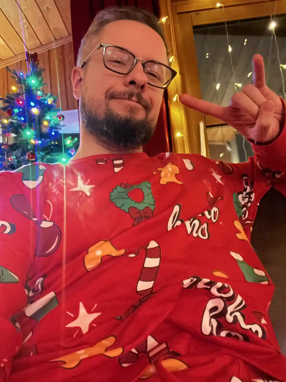 A man with glasses and a beard wears a red Christmas-themed sweater with candy canes, wreaths, and stars, posing with a…