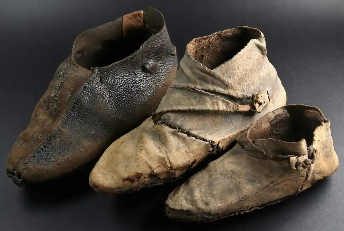 Three very old, worn, and tattered leather shoes are arranged on a dark surface. The shoes show significant signs of age and…