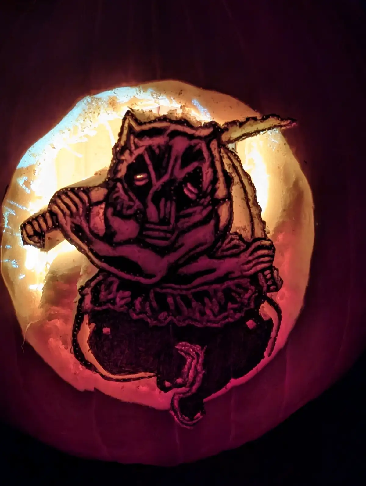 A carved pumpkin illuminated from within, featuring a menacing, stylized witch with pointed ears, holding a wand, and the…