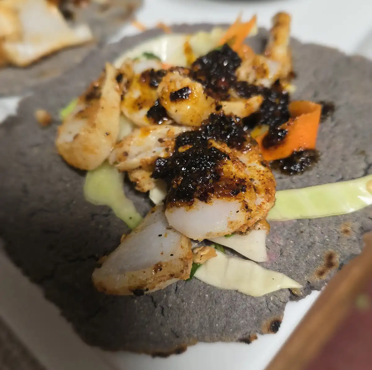A close-up photo of a dark blue corn tortilla topped with grilled shrimp, shredded cabbage, carrot strips, and a dark…