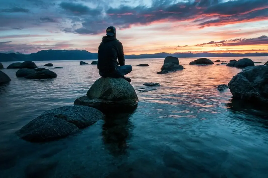 A person sits cross-legged on a large rock in calm water, facing a colorful sunset with mountains in the distance. The…
