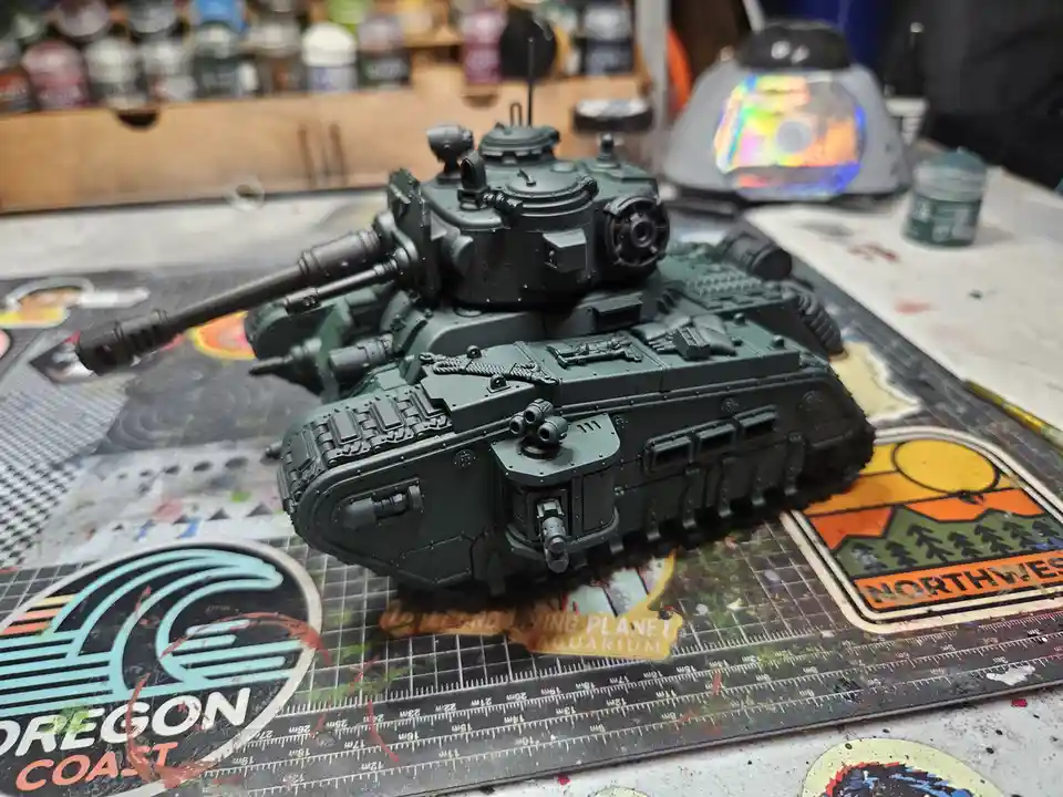 A detailed, dark green painted model tank sits on a cluttered hobbyist workbench with paint supplies, rulers, and stickers…