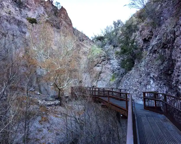 A photo of a metal walkway or bridge winding through a rocky canyon with bare trees and sparse vegetation. The walkway has…