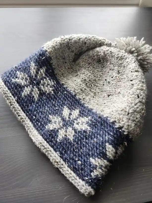 A hand-knitted beanie with a blue and white patterned section and a gray interior, featuring a pom-pom, rests on a dark…