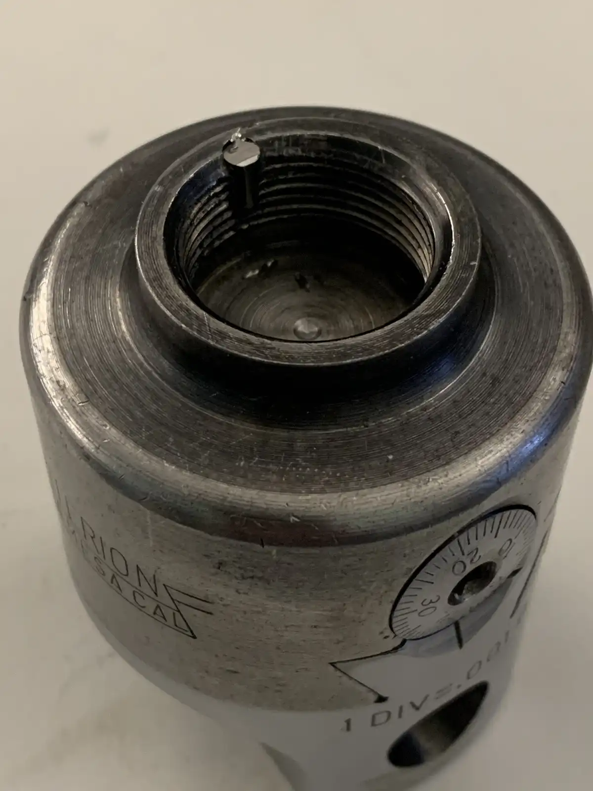 A close-up photo of a worn, metallic cylindrical gauge with visible threading and markings, including 'FERION MESACAL' and a…