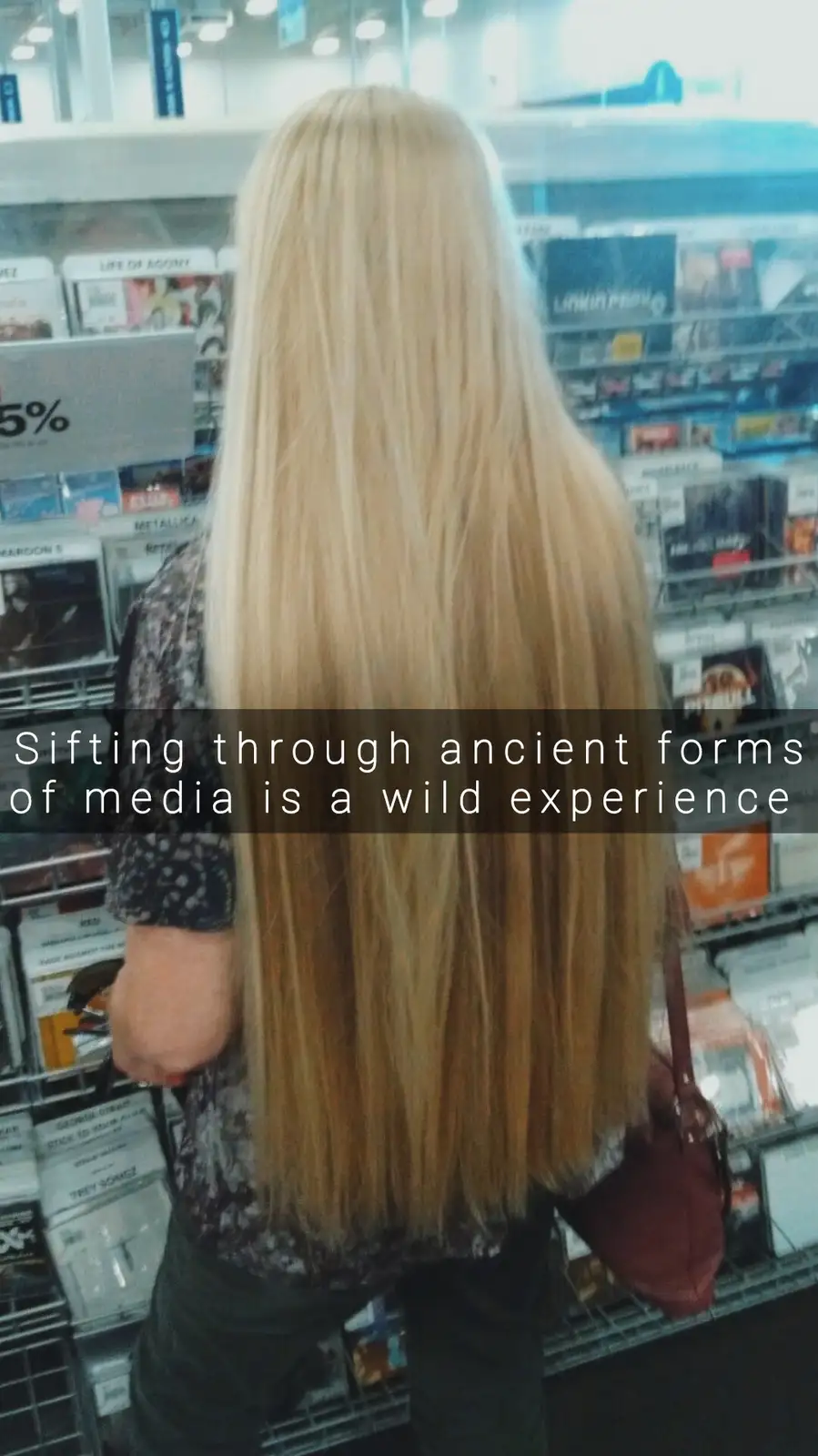 A person with long blonde hair viewed from behind stands in a store aisle filled with CD cases, their hair obscuring their…