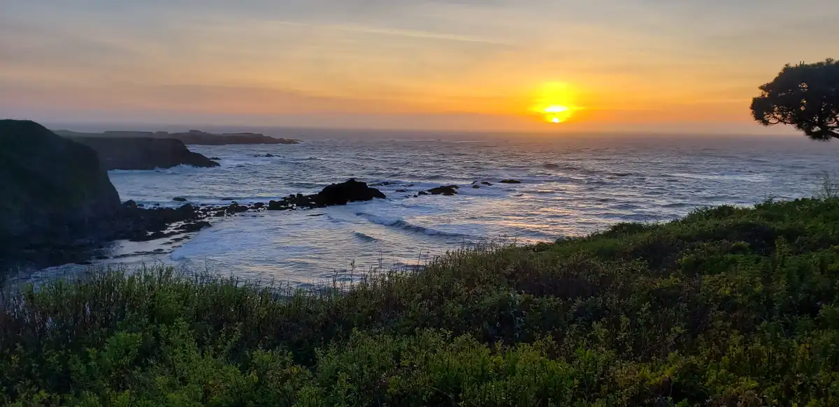 A scenic coastal sunset with the sun low on the horizon over the ocean, waves crashing against rocks, green shrubbery in the…
