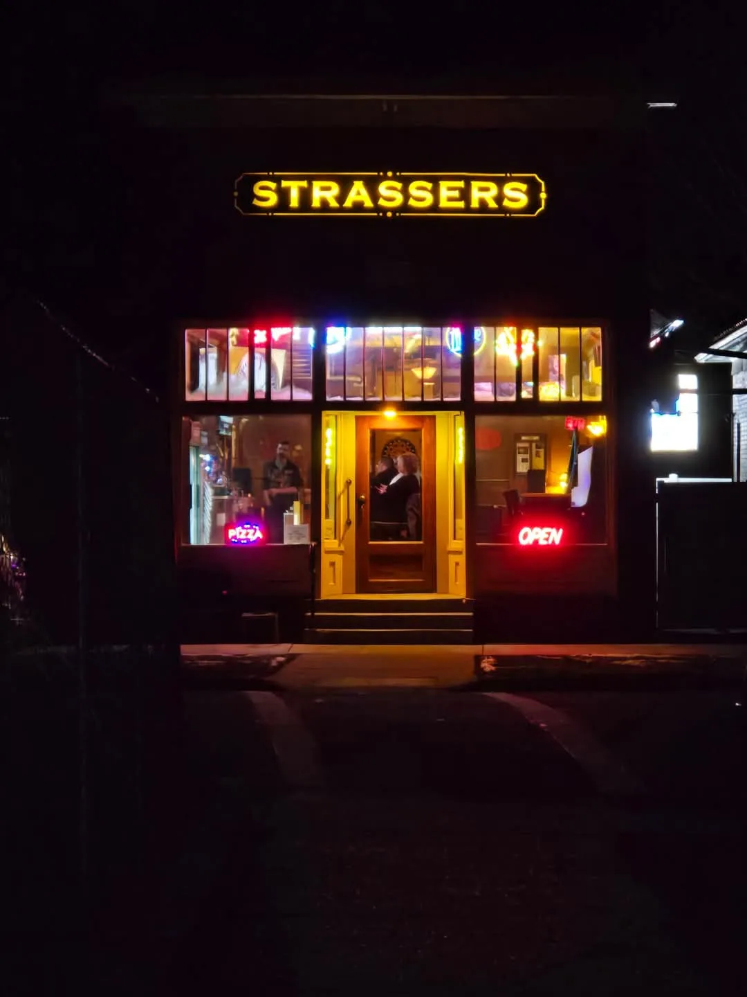A nighttime photo of a storefront with a glowing yellow neon sign reading 'STRASSERS' above the entrance. Inside, people are…