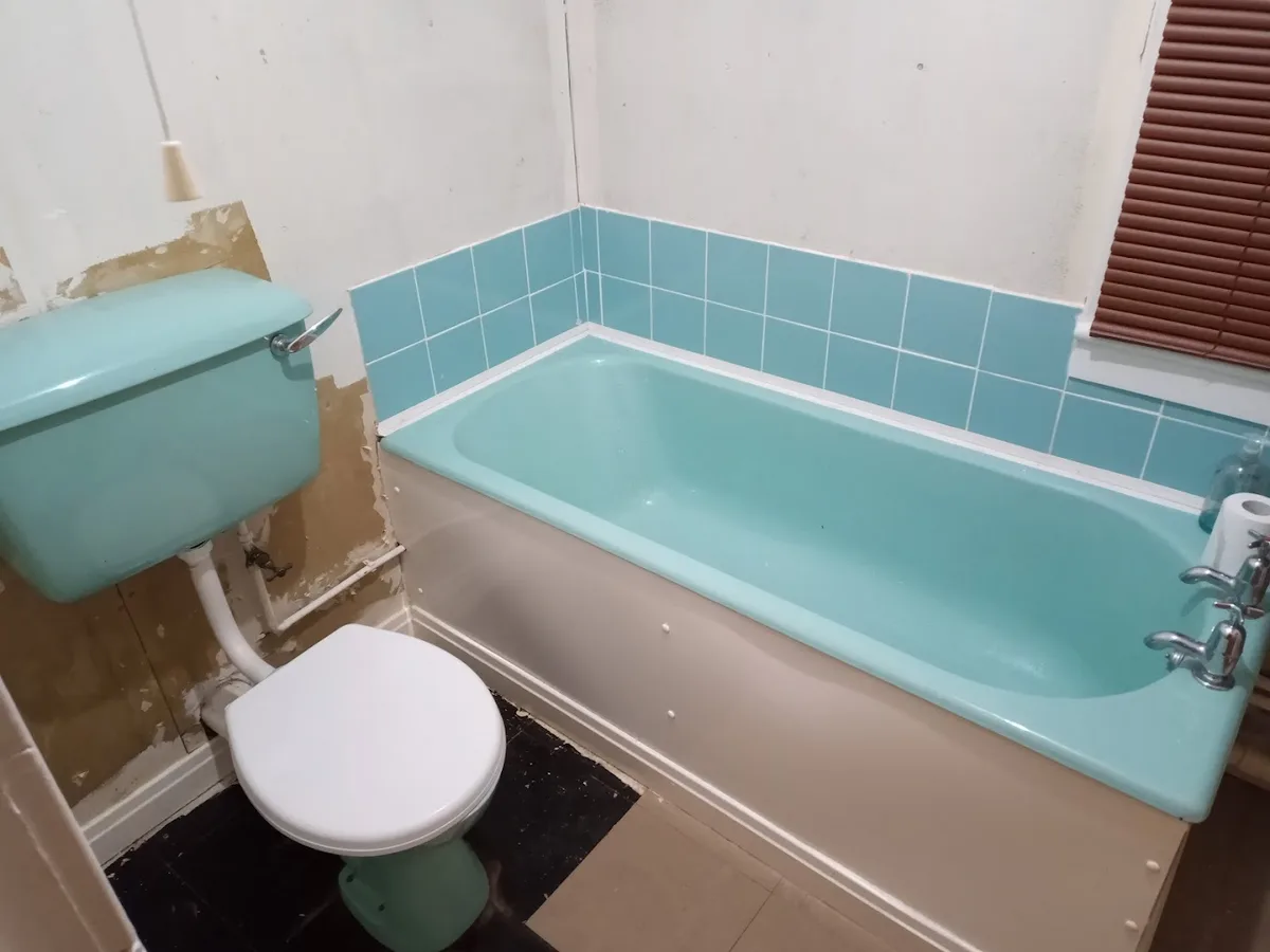 A bathroom with a turquoise bathtub and matching toilet, partially renovated with exposed drywall and pipes. The walls have…