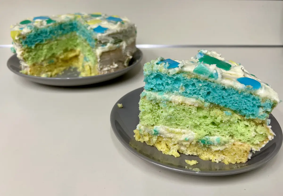 A slice of multi-layered cake with blue, green, and yellow sponge layers and white frosting sits on a gray plate in the…