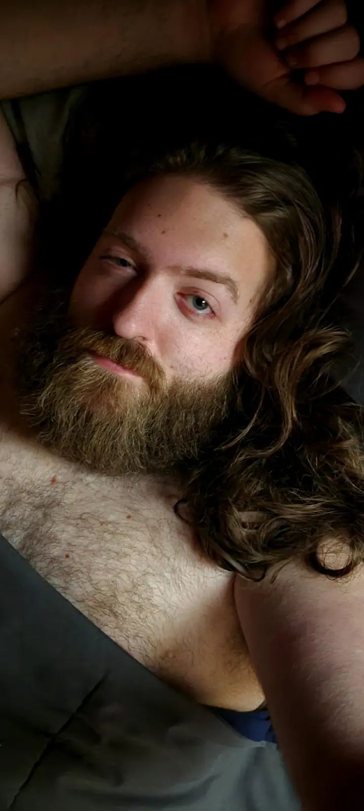 A shirtless man with long brown hair and a full beard lies in bed, looking directly at the camera with a relaxed, slightly…