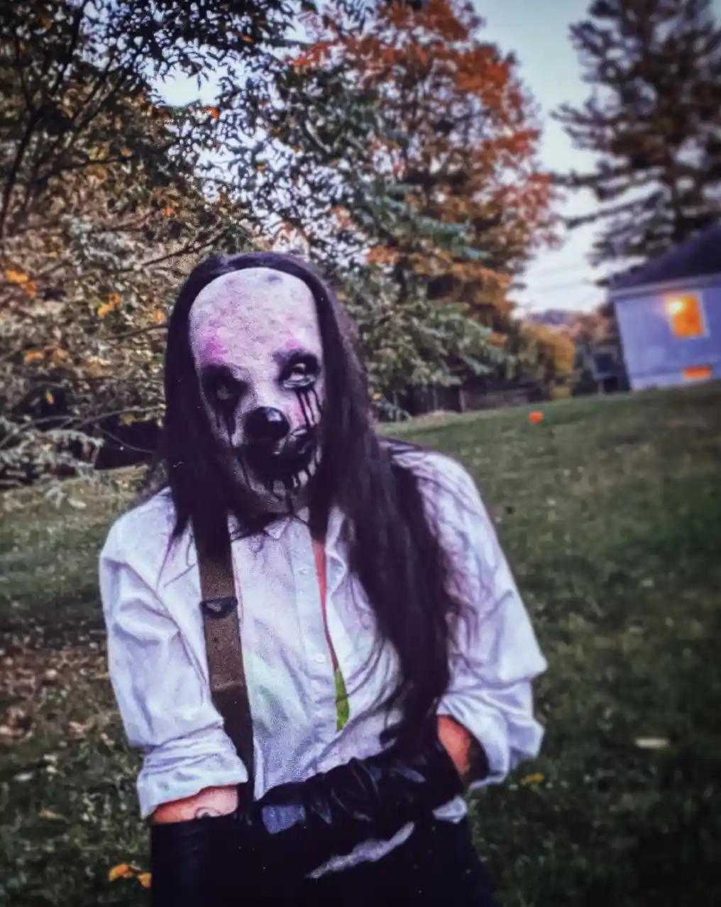 A person wearing a scary clown mask with black tears running down their face, long dark hair, a white button-up shirt, and…