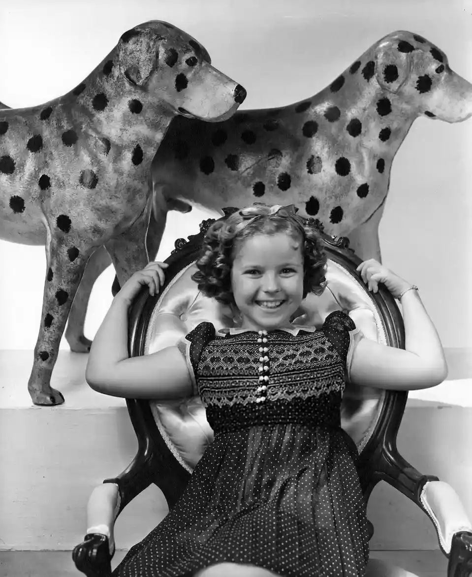 A vintage black-and-white photo shows a young girl in a polka-dot dress smiling while seated in an ornate chair, with two…